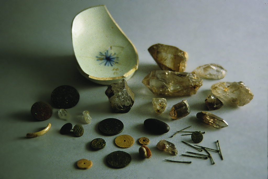 Objects from the Carroll House cache included shell discs, straight pins, buttons, two pierced coins, a tiny faceted glass bead, a smooth black stone, and large rock crystals. The collection was covered with an overturned pearlware bowl. Archaeology in Annapolis/ University of Maryland