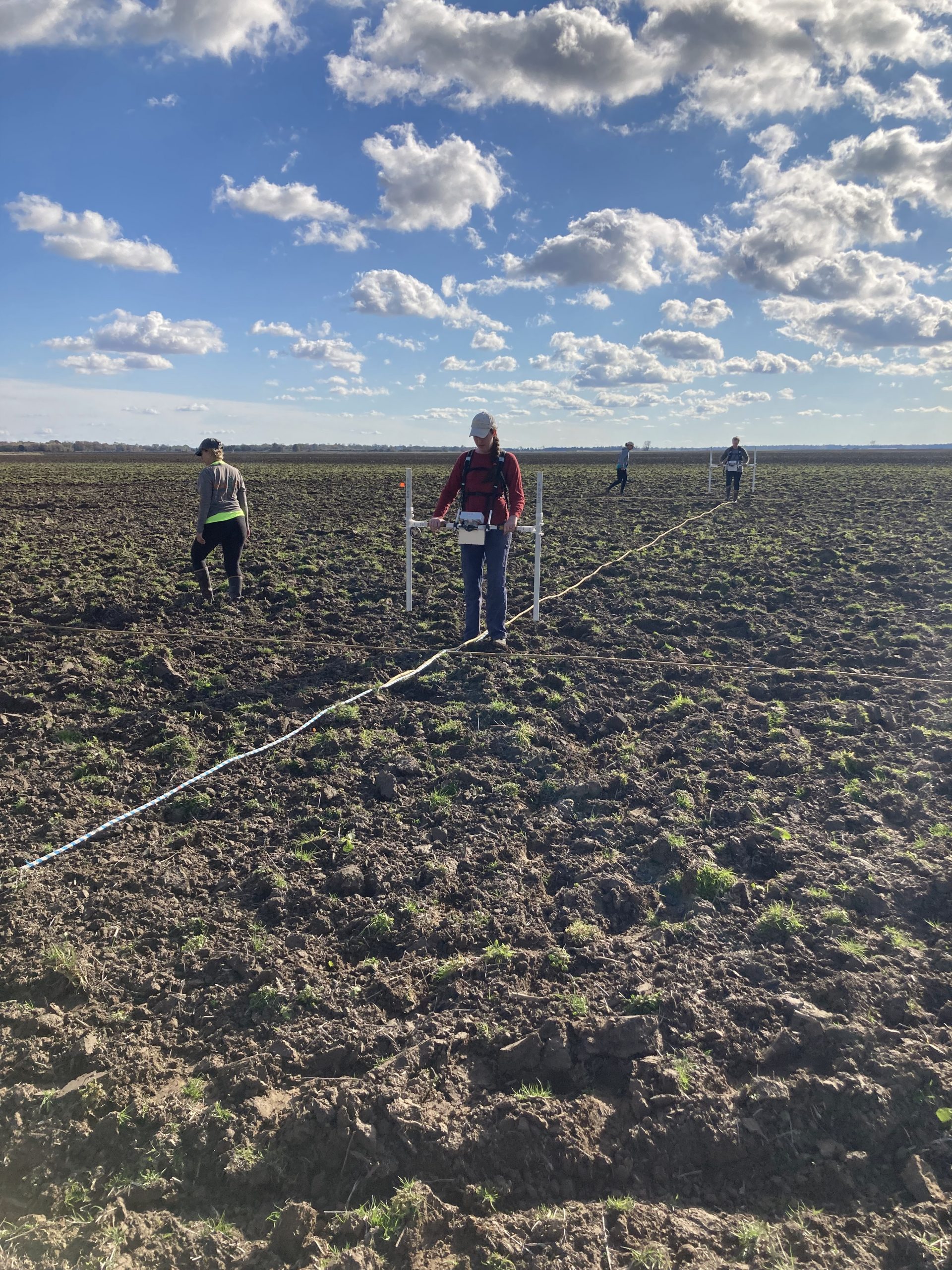 Photo of four people carrying equipment as they walk through a plowed field.