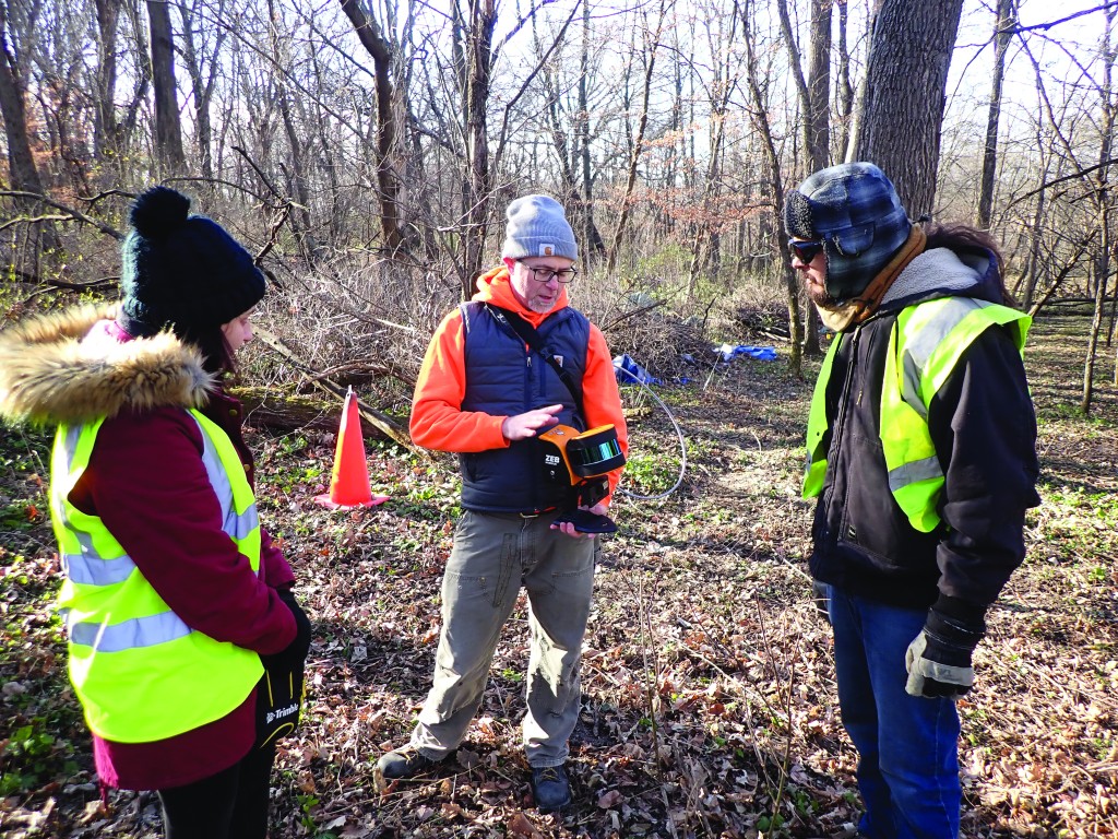 photo of three people standing in a wooded area holding a device. an orange cone is in the background.