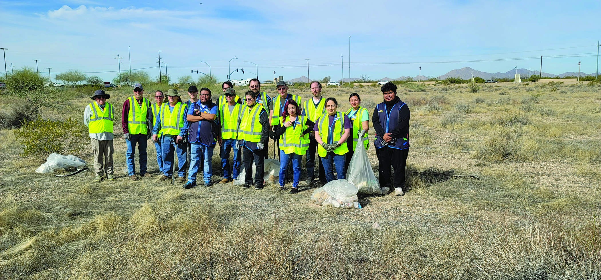 photo of a group of approximately 15 people who are standing in a field, wearing bright yellow caution vests and holding trashbags.