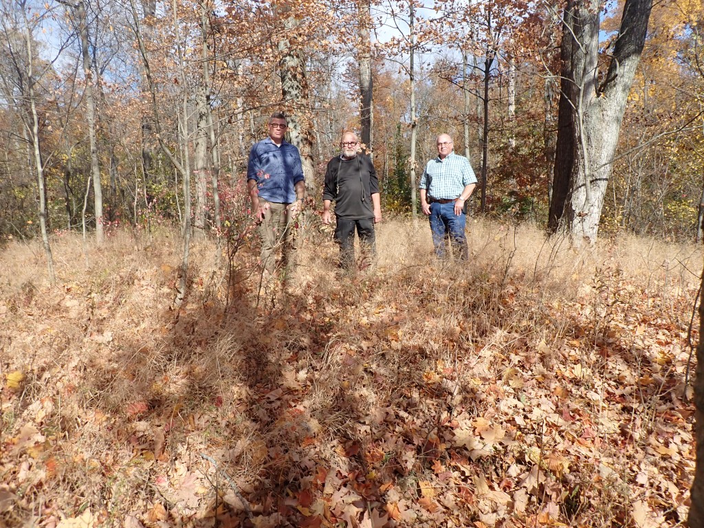 Photo of 3 men standing on leaf and brush covered mound with deciduous trees in the background.