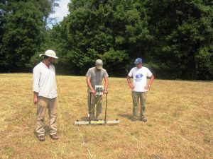 photo of three men with gradiometry tools standing in a recently mowed field surrounded by trees.